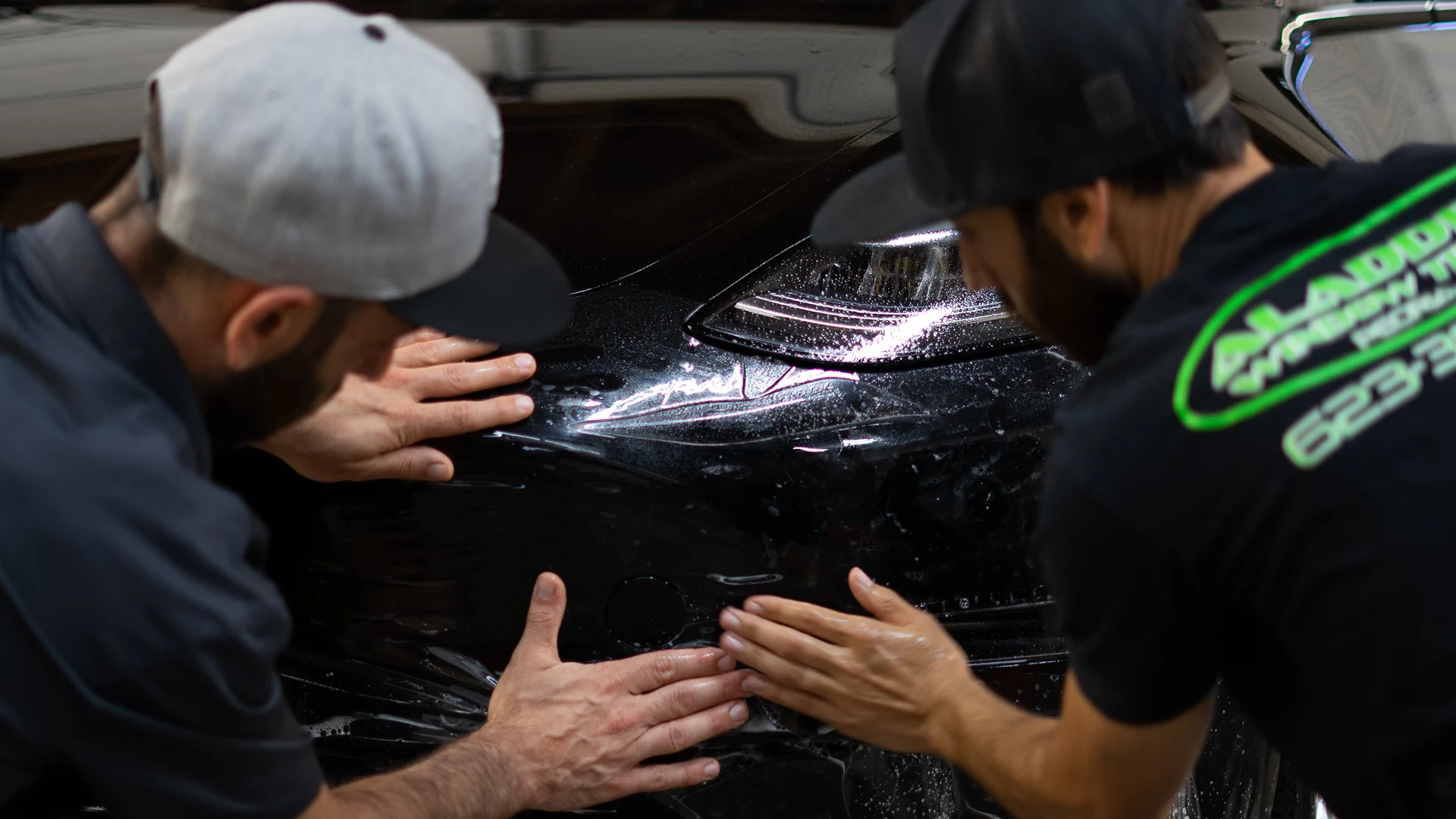 Two Aladdins Window Tinting installers applying clear paint protection film to a black vehicle’s front bumper.