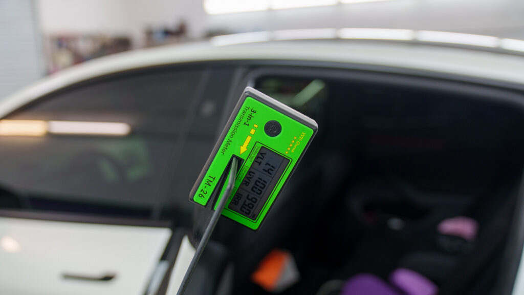 Digital tint meter measuring visible light transmission through a white vehicle’s tinted side window.