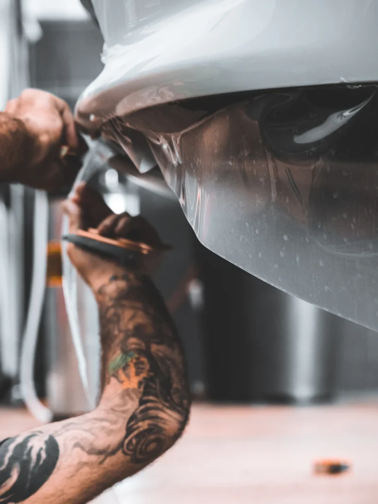 Close-up of an installer smoothing clear paint protection film around a white vehicle’s lower bumper with a squeegee.