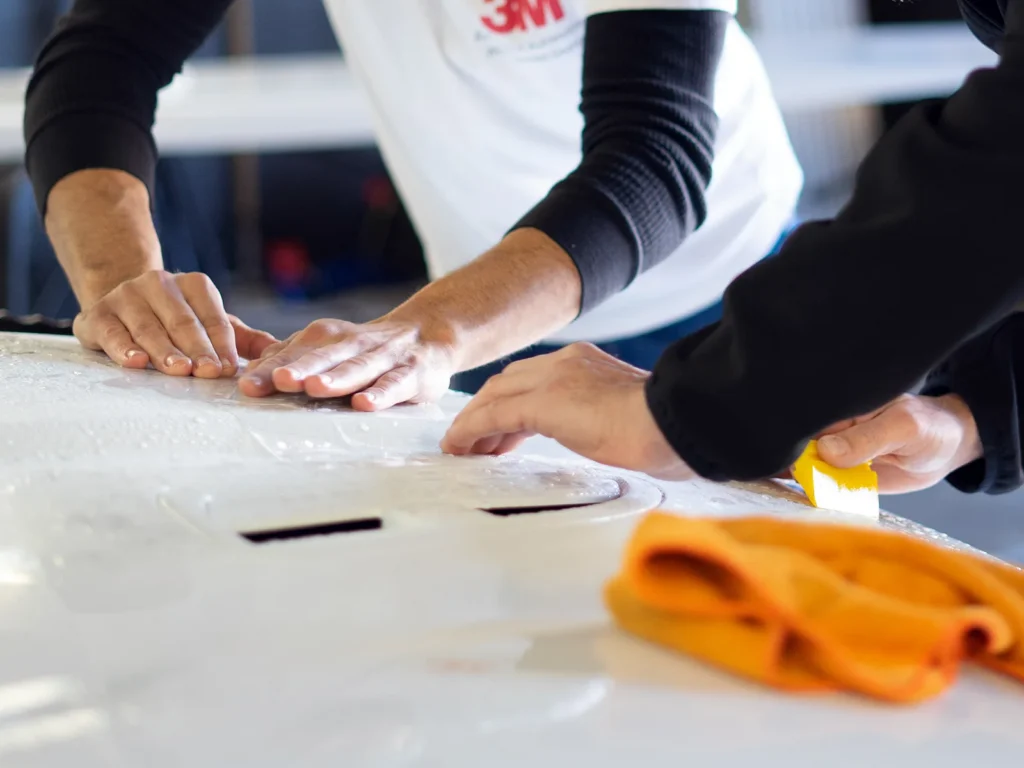 Installers applying clear paint protection film to a white vehicle’s hood with a squeegee.