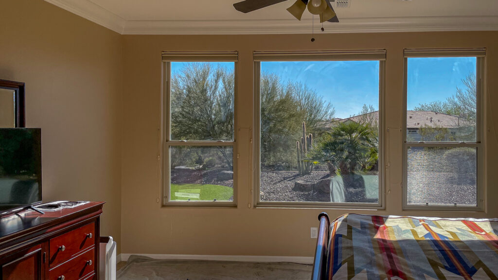 Living room with three large windows overlooking a sunny backyard with trees and desert landscaping.