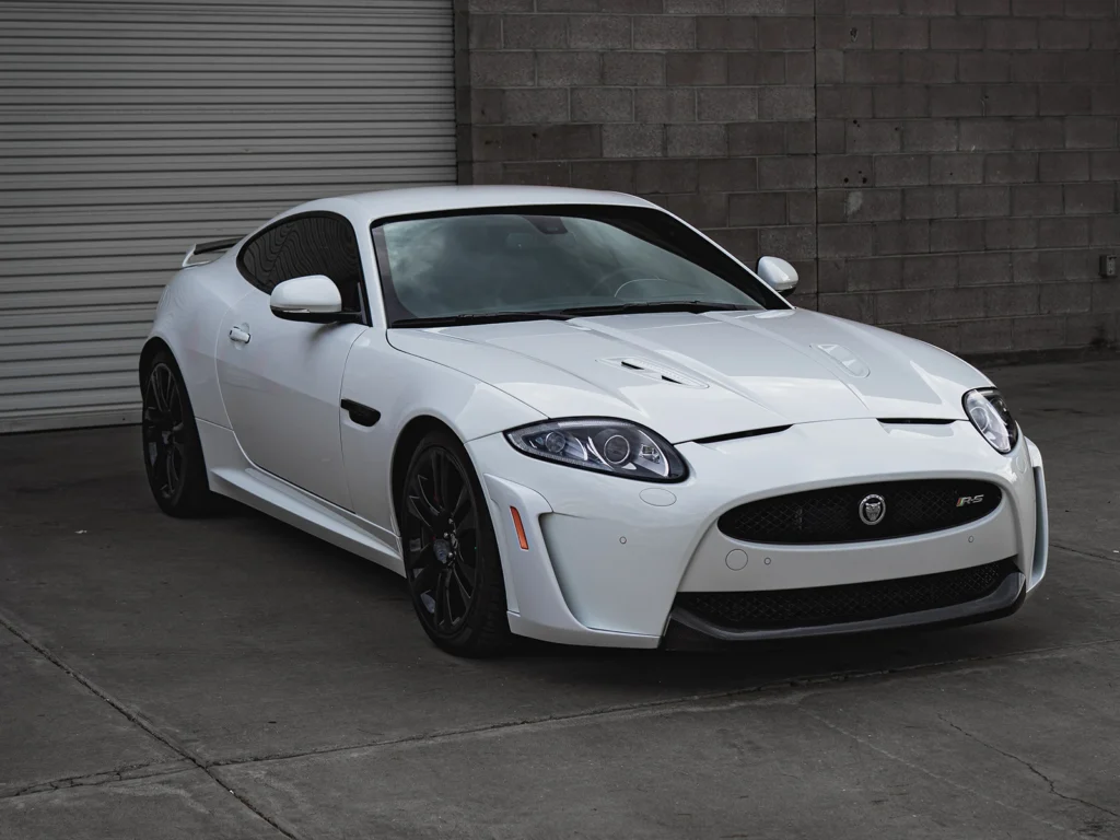 White Jaguar XKR-S with black wheels parked outside the shop, viewed from the front passenger side.